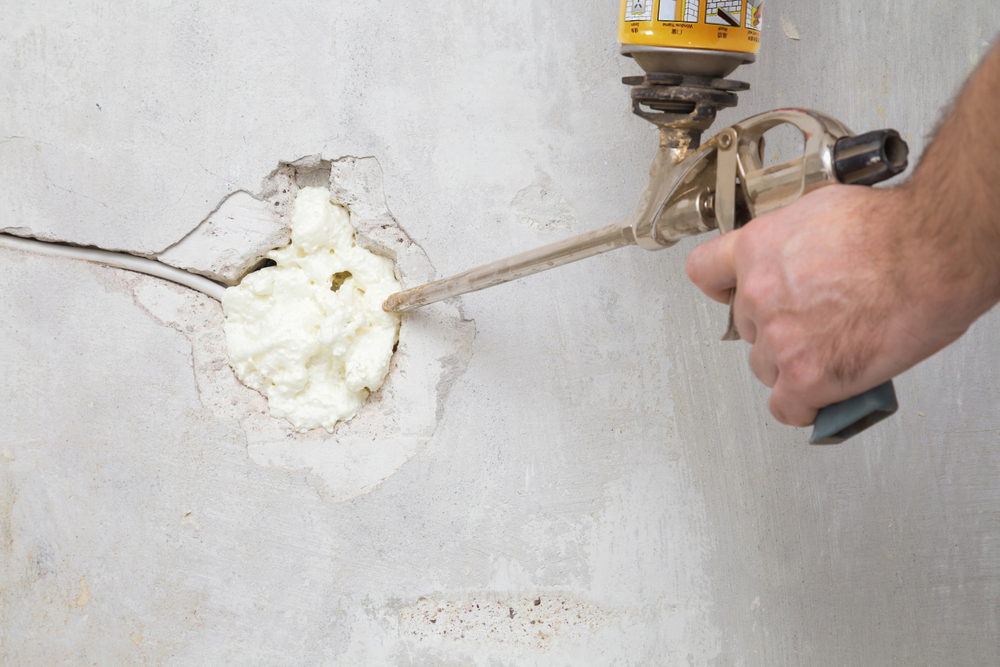 Young man hand using spray gun and filling hole with construction foam in concrete wall where was old outlet. Closeup. Repair work of home. Renovation process. Side view.